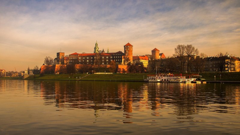 Wawel o zachodzie słońca – panorama królewskiego zamku w Krakowie z ciepłymi barwami nieba odbijającymi się w Wiśle.